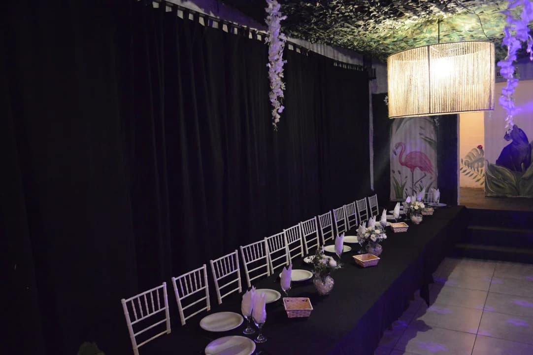 Mesa imperial larga con sillas Tiffany blancas, servilletas en copa, centros de mesa florales y mural de flamenco rosa al fondo contra cortinado negro