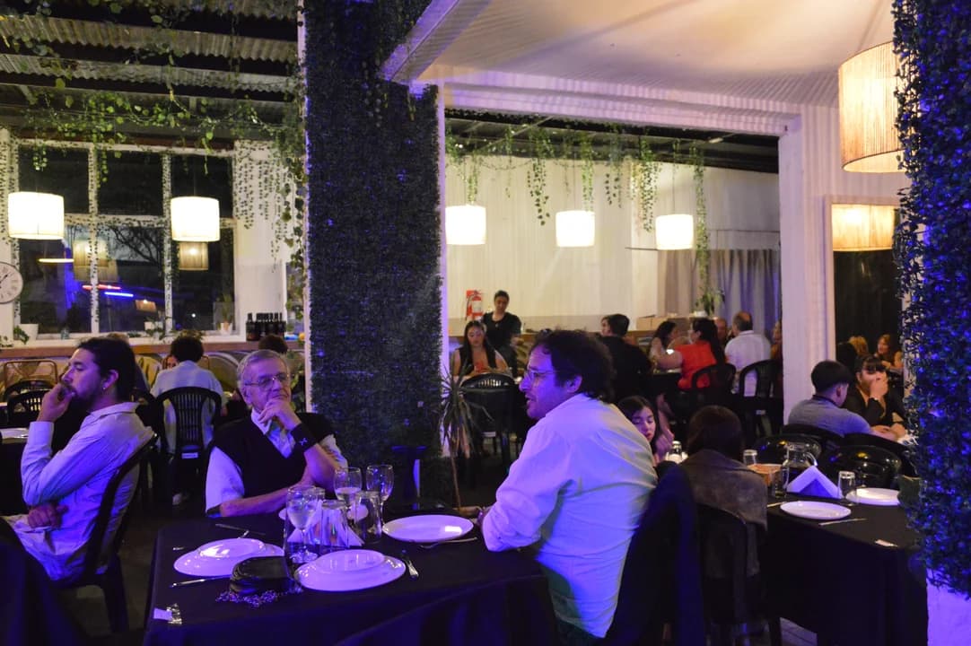Salón principal en plena celebración con columnas decoradas con follaje, invitados cenando y cortinas blancas dividiendo ambientes bajo iluminación cálida y violeta