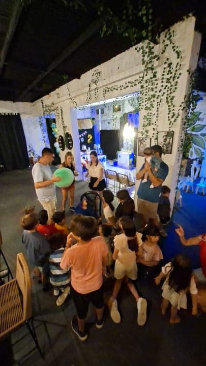 Fiesta infantil con niños reunidos y animador con pelota, plantas colgantes y luces azules