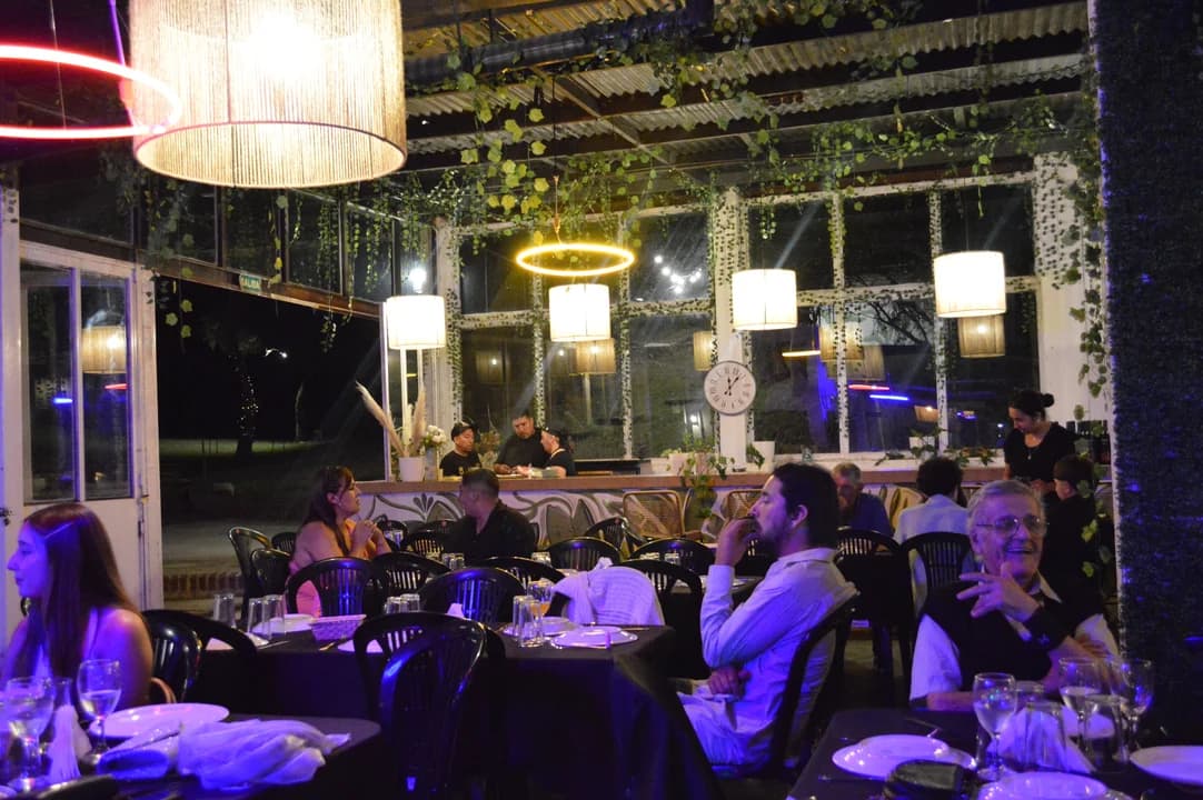 Vista panorámica del salón durante una cena: invitados en sus mesas bajo lámparas cilíndricas, aros LED dorados y enredaderas colgantes con ventanales al jardín nocturno