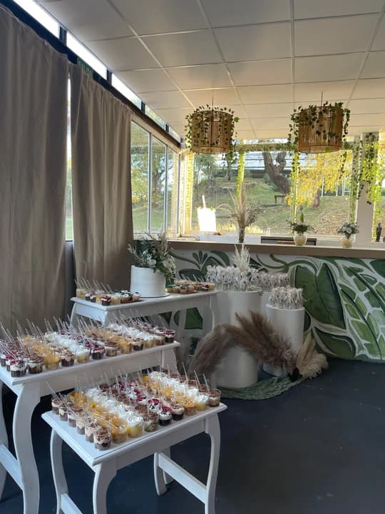 Mesa dulce elegante con vasitos de postres, torta blanca, pampas y murales de hojas