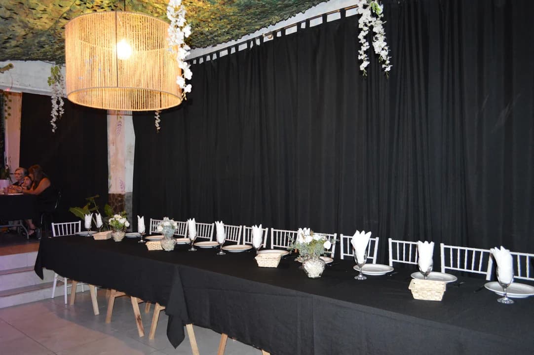 Mesa imperial completa vista desde el costado con sillas Tiffany, centros florales blancos, lámpara de mimbre colgante y guirnaldas de flores sobre cortinado negro